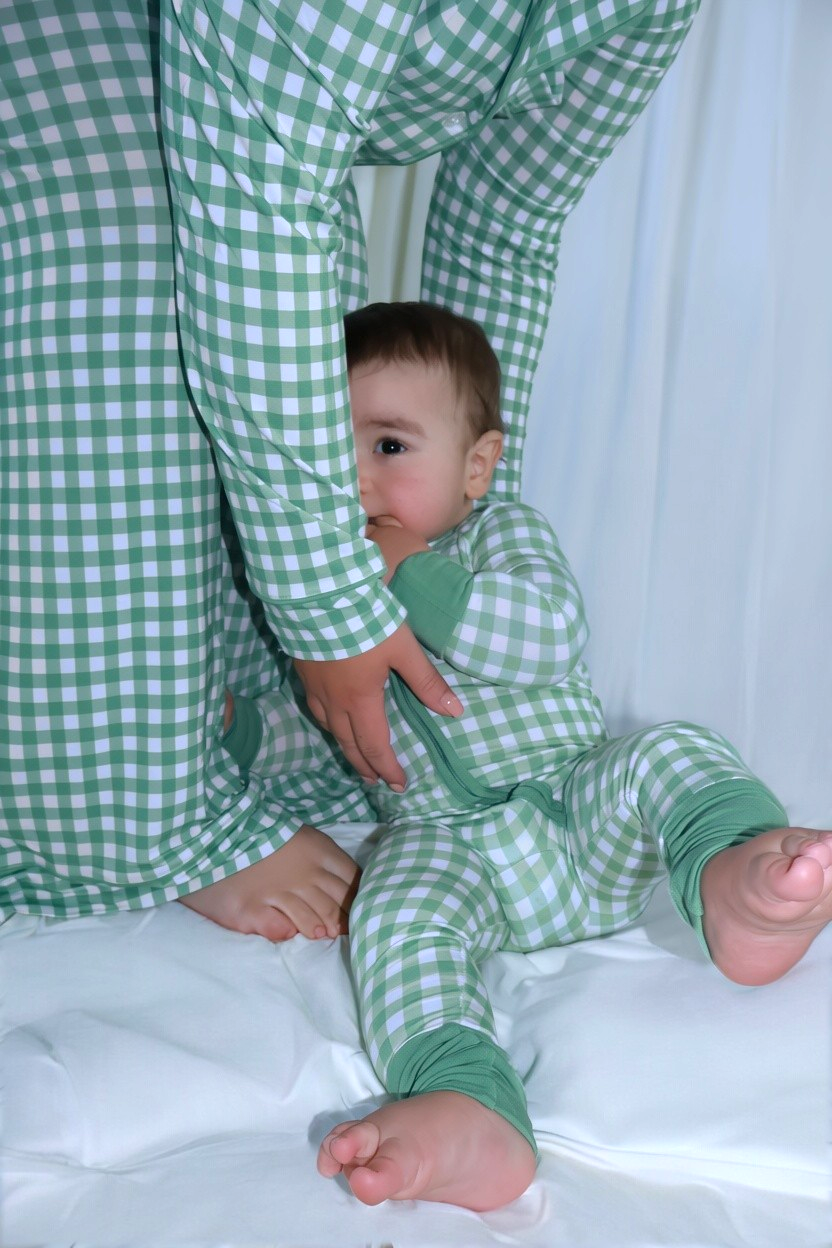 Baby seated at mom's feet on white bedding, both wearing matching Goodnight Goose green gingham bamboo pajamas.